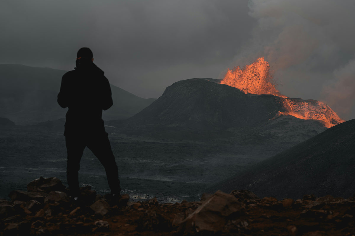 A silhouetted person stands on a rocky landscape looking toward a dark mountain with bright orange lava or fire glowing on the slope under a smoky gray sky.